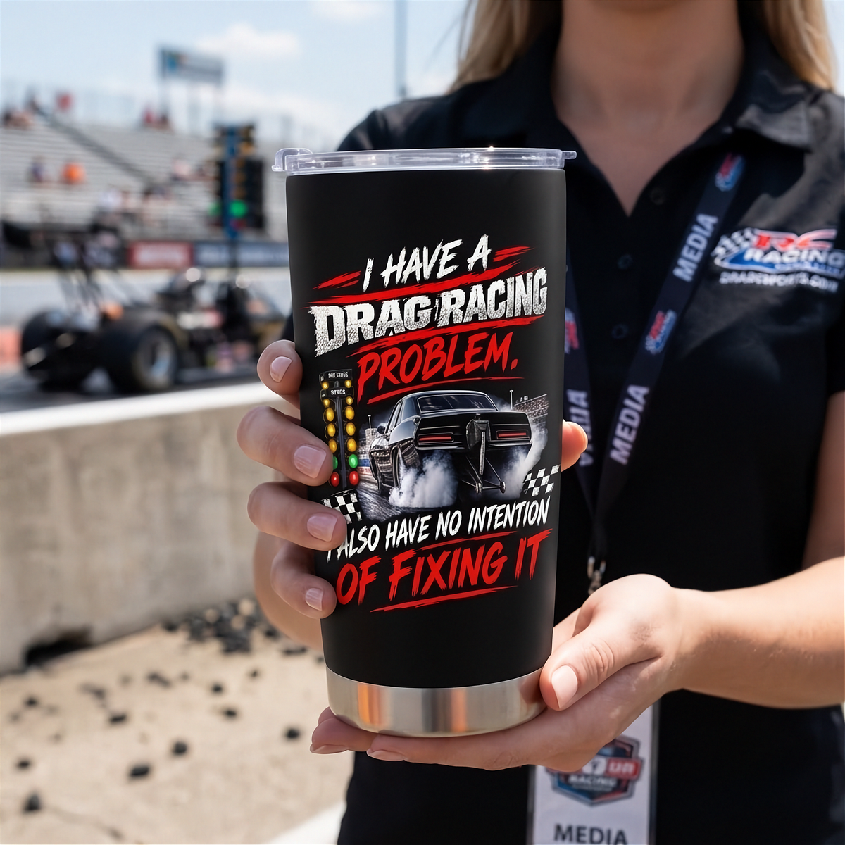 A person in a black racing media shirt holds a black insulated tumbler with a clear lid. The tumbler has a laser-engraved design featuring a drag racing car, checkered flags, and the text 'I HAVE A DRAG RACING PROBLEM, I ALSO HAVE NO INTENTION OF FIXING IT' in white and red lettering.