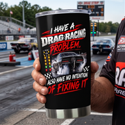A person holds a black insulated tumbler with a clear lid at a drag racing track. The tumbler features a graphic of a drag racer car and the text 'I HAVE A DRAG RACING PROBLEM.