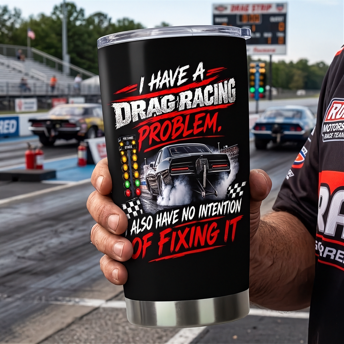 A person holds a black insulated tumbler with a clear lid at a drag racing track. The tumbler features a graphic of a drag racer car and the text 'I HAVE A DRAG RACING PROBLEM.