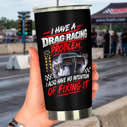 A hand holds a black insulated tumbler with a silver rim and base. The tumbler features a graphic of a drag racing car and the text 'I HAVE A DRAG RACING PROBLEM.