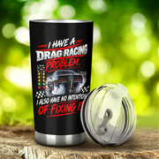 A black insulated tumbler with a clear lid sits on a wooden surface against a blurred green outdoor background. The tumbler has a drag racing graphic with the text 'I HAVE A DRAG RACING PROBLEM, I ALSO HAVE NO INTENTION OF FIXING IT' and a car illustration.