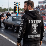 A man stands on a drag racing track wearing a black hoodie with a white graphic print on the back that reads 'I STRIP ON WEEKENDS' and 'DRAG RACING' printed down both sleeves. A black muscle car is visible in the drag lane behind him, with race lights and a crowd in the background.
