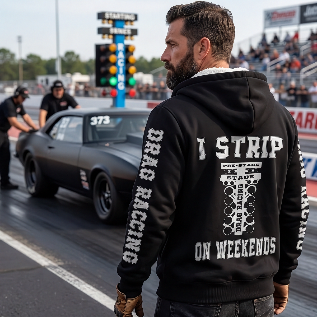 A man stands on a drag racing track wearing a black hoodie with a white graphic print on the back that reads 'I STRIP ON WEEKENDS' and 'DRAG RACING' printed down both sleeves. A black muscle car is visible in the drag lane behind him, with race lights and a crowd in the background.