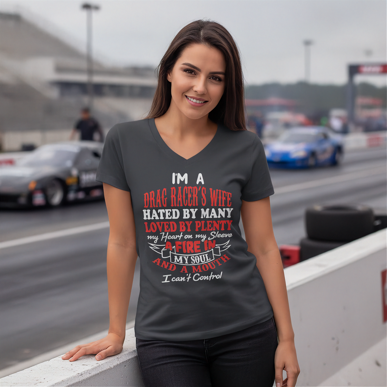 A woman stands on a racetrack wearing a dark gray V-neck t-shirt with a humorous drag racer-themed graphic print. The background shows blurred race cars and track barriers.