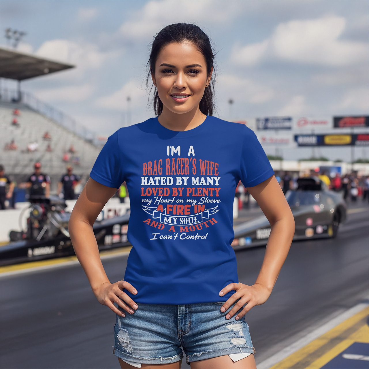 A woman stands on a racetrack wearing a royal blue graphic t-shirt with a humorous drag racer quote, paired with distressed denim shorts. The background shows blurred race cars and grandstands.