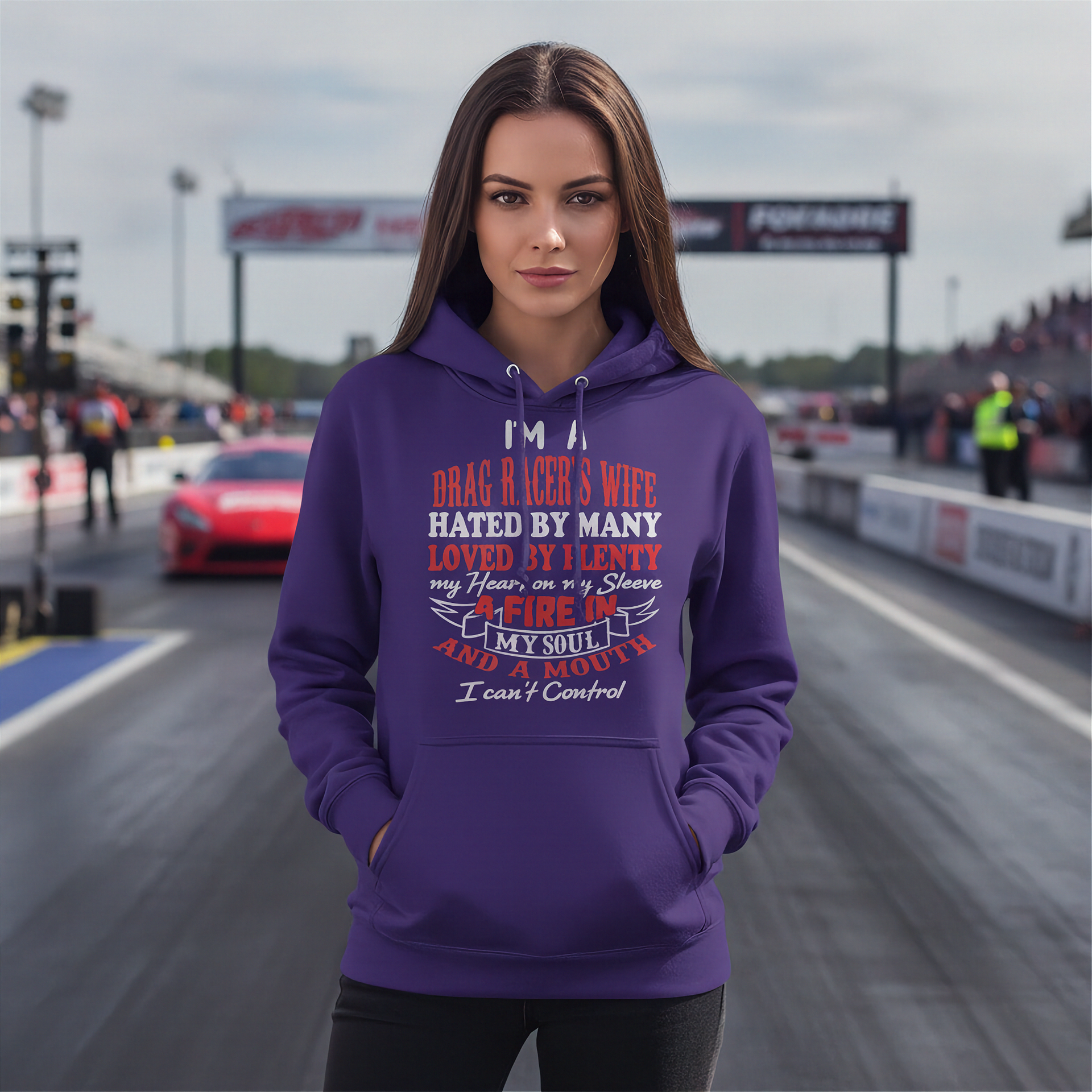 A woman stands on a drag racing track wearing a purple hoodie with a graphic text print on the front. The text reads: 'I'M A DRAG RACE WIFE HATED BY MANY LOVED BY MANY my heart is in a race AFIRE IN MY SOUL I can't Control'.