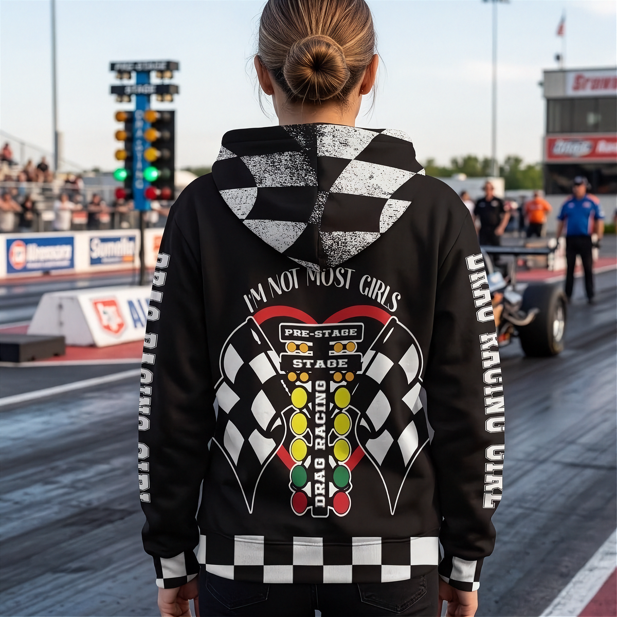 A person standing on a racetrack wearing a black hoodie with a large drag racing graphic on the back, featuring a checkered pattern, race flags, and the text 'I'M NOT MOST GIRLS' and 'DRAG RACING'.