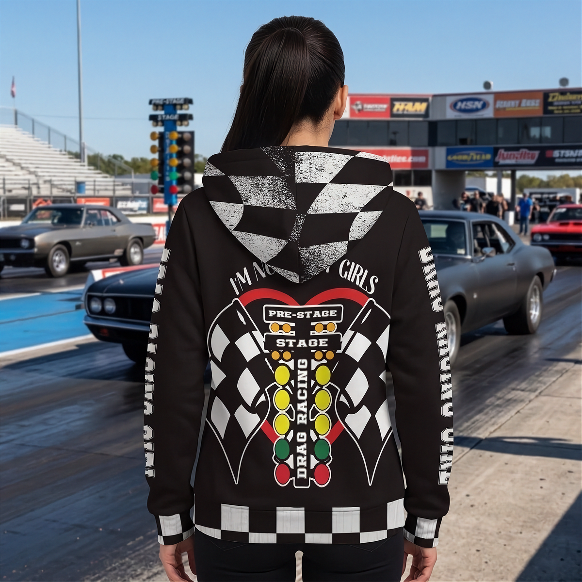 A woman viewed from behind wearing a black hoodie with a large drag racing graphic on the back, standing on a racetrack with drag cars and a building in the background.
