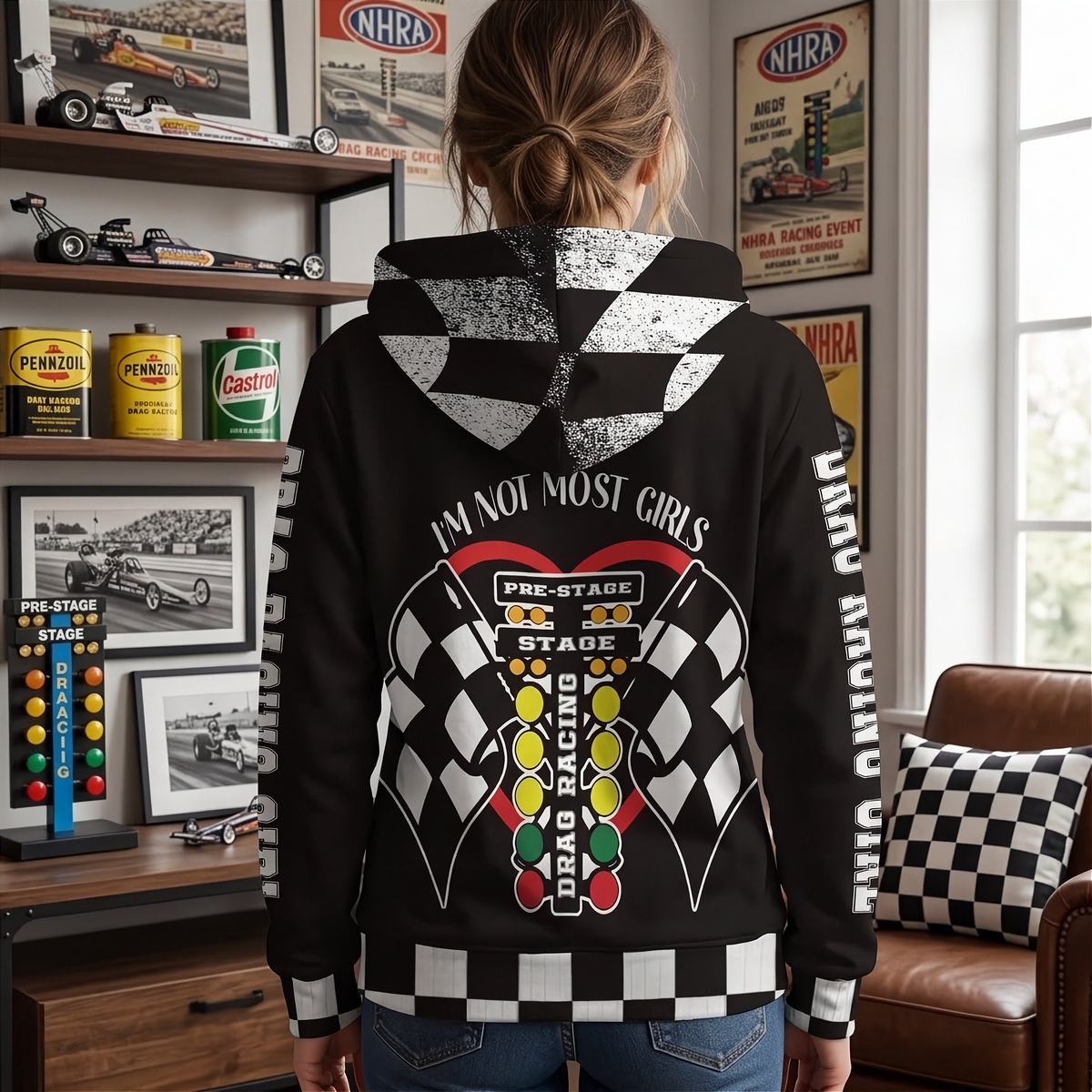 A woman shown from behind wearing a black hoodie with a large drag racing graphic on the back, standing in a room with racing memorabilia and a checkered seat cushion.