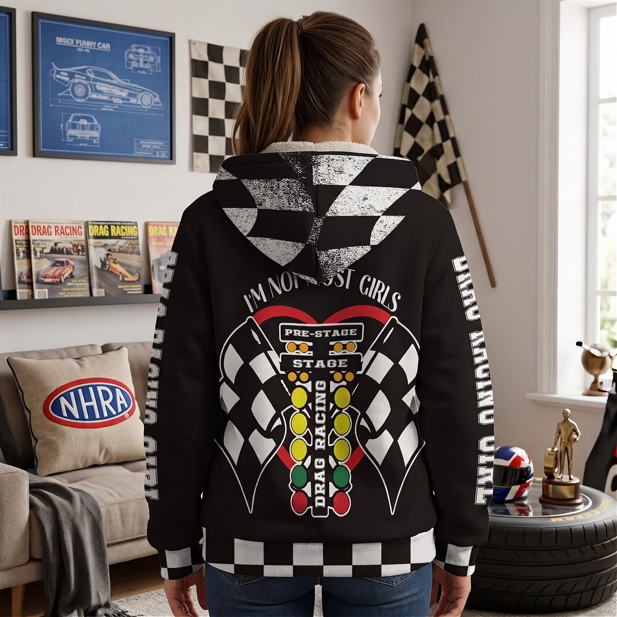 A woman shown from behind wearing a black hoodie with a detailed drag racing graphic on the back and checkered hood lining, standing in a decorated room with racing memorabilia.