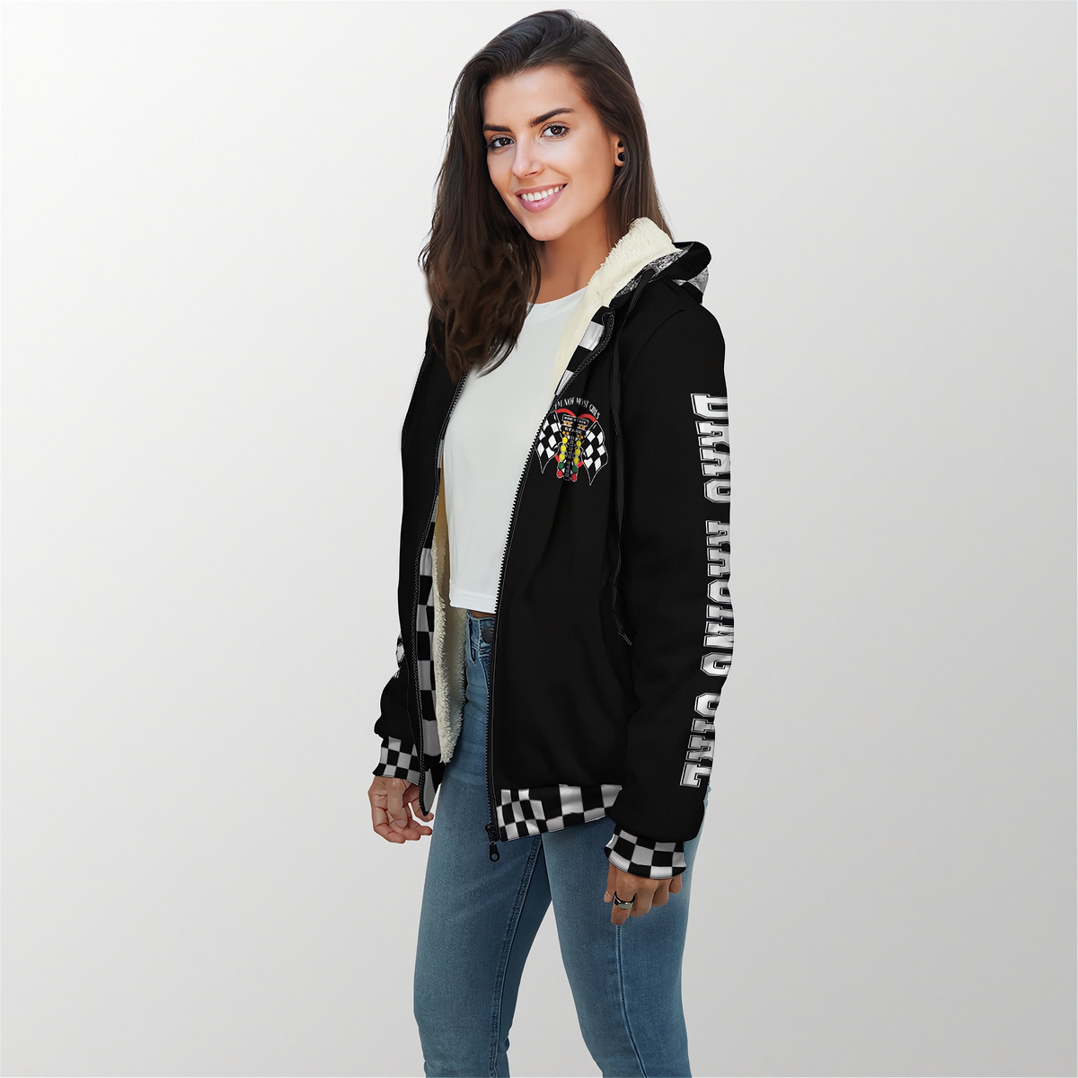 A woman modeling a black zip-up hoodie with a cream sherpa-lined hood, black-and-white checkered trim, and vertical white text on the sleeve, paired with a white top and blue jeans against a light gray studio background.