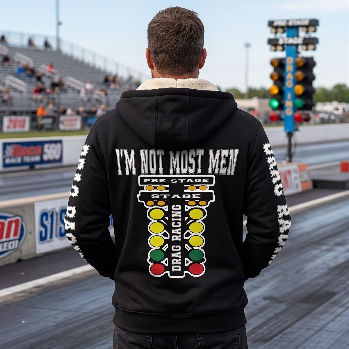 A man viewed from behind wearing a black hoodie on a drag racing track. The hoodie features a large graphic on the back with the text 'I'M NOT MOST MEN' and 'PRE-STAGE X STAGE DRAG RACING' above a vertical traffic-light-style graphic in red, yellow, and green.