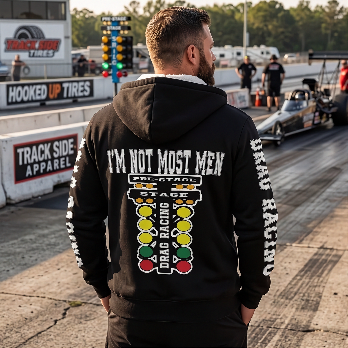 A man stands on a drag racing track wearing a black hoodie with a large racing-themed graphic on the back and 'DRAG RACING' printed down the sleeve. Drag cars and trackside signage are visible in the background.