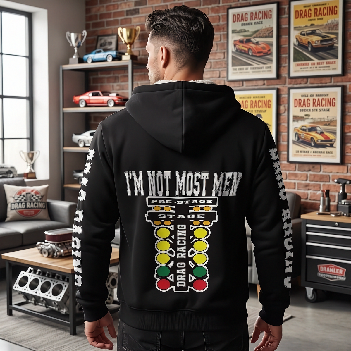 A man viewed from behind wearing a black hoodie with a large drag racing graphic and the text 'I'M NOT MOST MEN' on the back, standing in a garage-themed room with posters and shelves in the background.