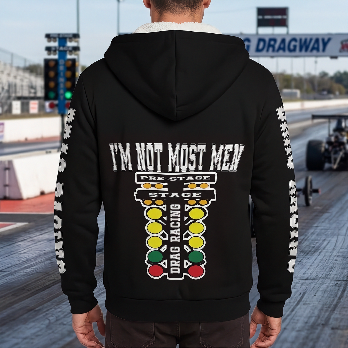 A man stands on a drag strip wearing a black hoodie from the back, featuring a large graphic with the text 'I'M NOT MOST MEN' and a drag racing theme. The hoodie has white text down both sleeves and a colorful graphic in the center.