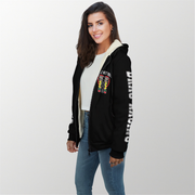 A woman modeling a black zip-up hoodie with a cream sherpa lining, featuring a colorful graphic on the left chest and white text down the right sleeve, paired with a white t-shirt and blue jeans against a light gray background.