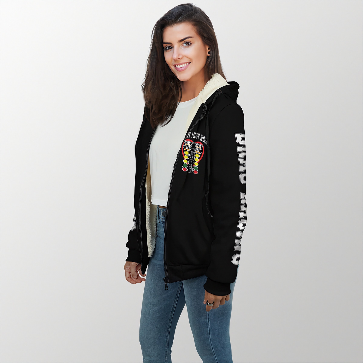 A woman modeling a black zip-up hoodie with a cream sherpa lining, featuring a colorful graphic on the left chest and white text down the right sleeve, paired with a white t-shirt and blue jeans against a light gray background.