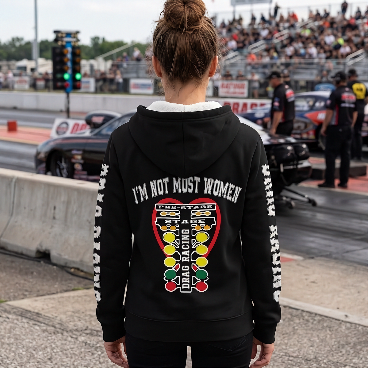 A woman viewed from behind wearing a black hoodie with a large graphic on the back and white text down the sleeves, standing at a racetrack with cars and a crowd in the background.