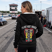 A woman stands on a racetrack wearing a black hoodie with a large drag racing graphic on the back and 'I'M NOT MOST WOMEN' printed across the upper back. The scene includes race cars, trailers, and track facilities in the background.