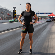 A woman stands on a drag racing track wearing a black short-sleeve t-shirt and black shorts with a colorful ''I'M NOT MOST WOMEN DRAG RACING'' pouch attached to the waist. The background shows the racetrack, grandstands, and race cars.