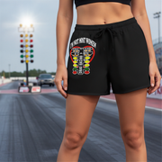 A person wearing black athletic shorts with a large 'I'M NOT MOST WOMEN DRAG RACING' graphic print on the front, standing on a racetrack. The shorts have a drawstring waist and the graphic features a red heart, yellow and green elements, and white text reading 'I'M NOT MOST WOMEN'.