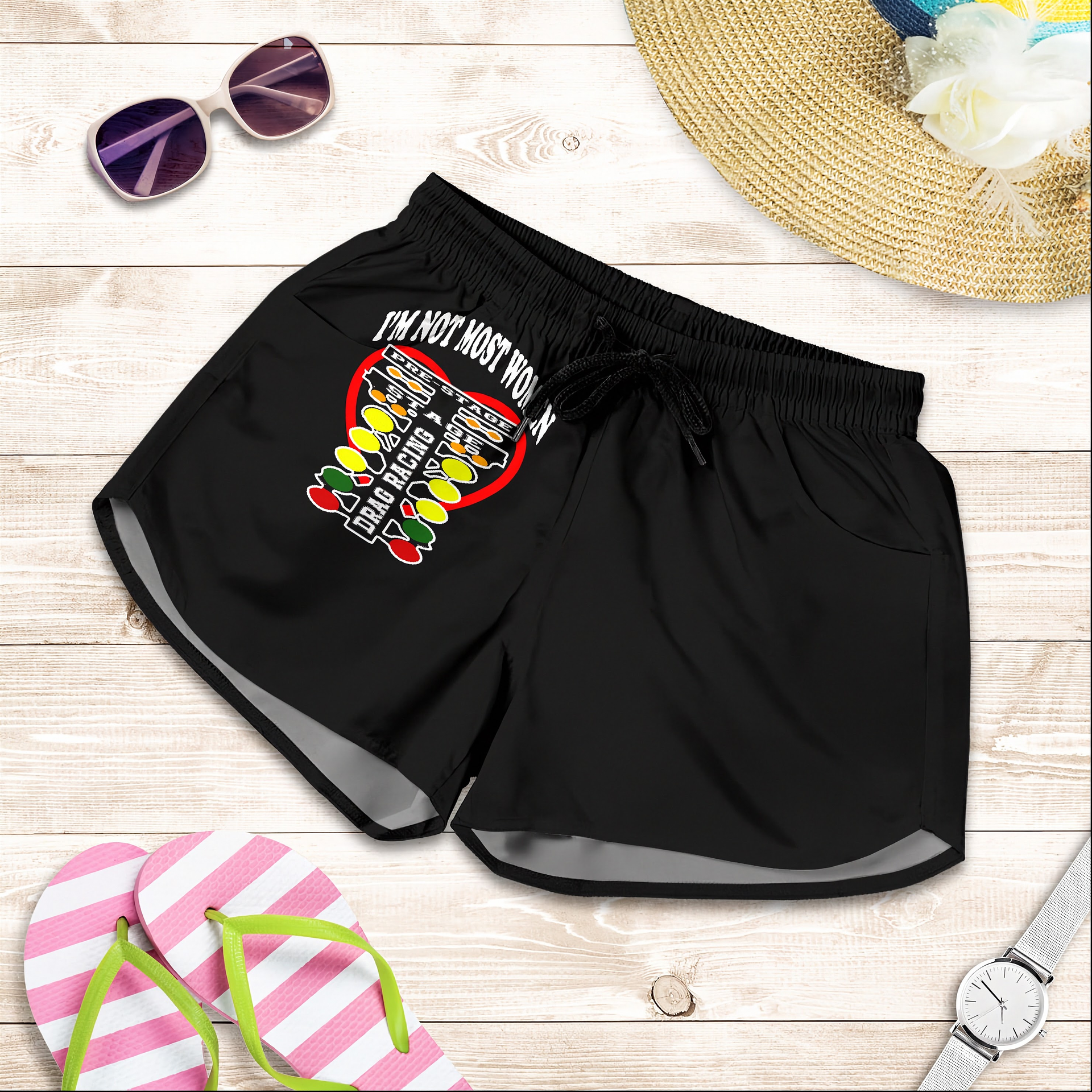 Black athletic shorts with a 'I'M NOT MOST WOMEN DRAG RACING' graphic and text on the left leg, styled in a flat lay with pink-and-white striped flip-flops, sunglasses, a straw hat, a watch, and a flower on a light wood background.