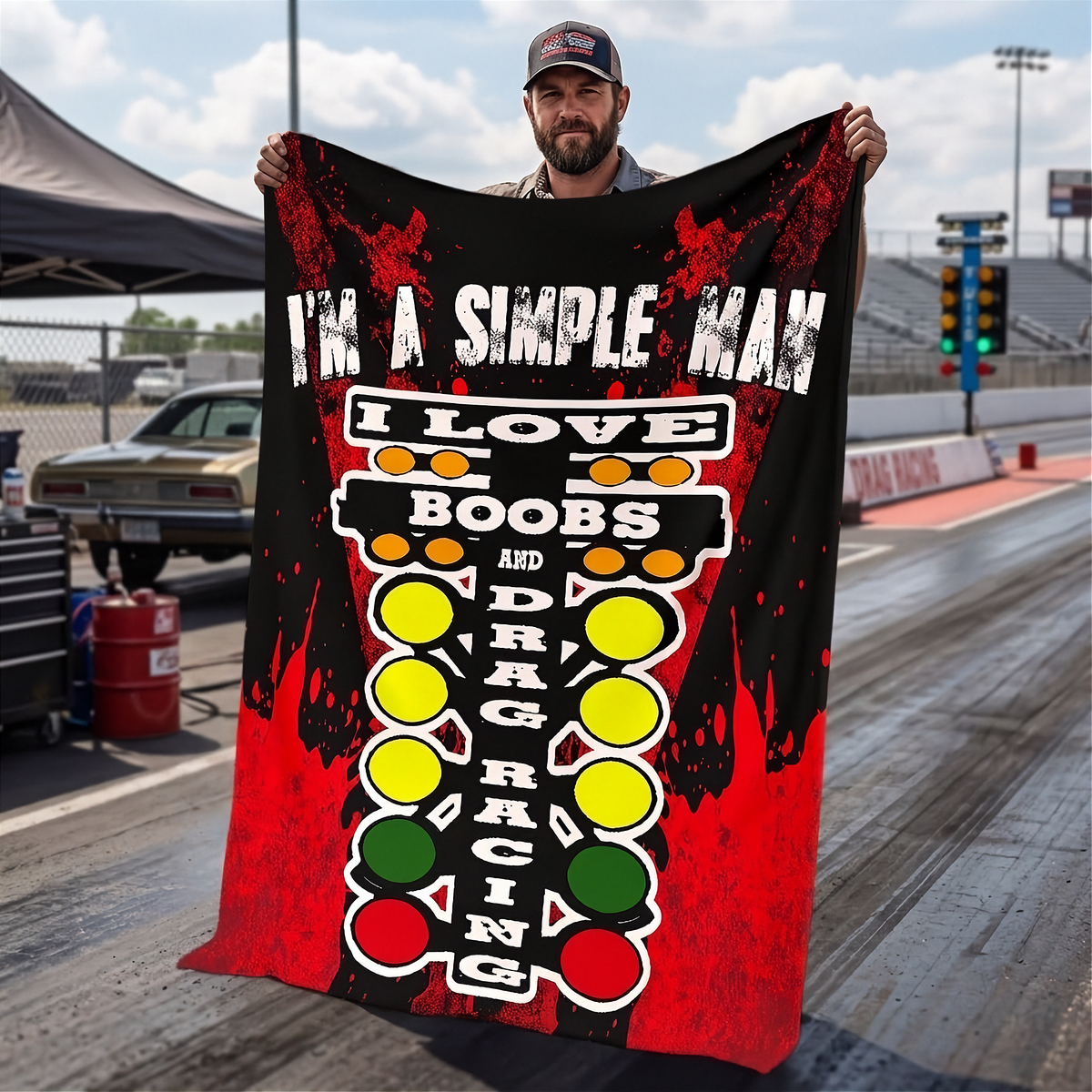 A man standing on a drag strip holds up a large black and red graphic blanket featuring the text 'I'M A SIMPLE MAN I LOVE BOOBS AND DRAG RACING' with yellow, green, and white racing light illustrations.