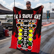 A man standing on a drag strip holds up a large black and red graphic blanket featuring the text 'I'M A SIMPLE MAN I LOVE BOOBS AND DRAG RACING' with yellow, green, and white racing light illustrations.