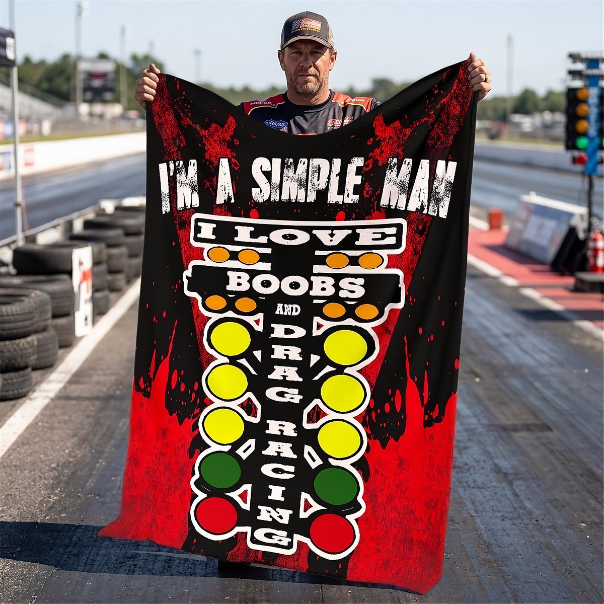 A man standing on a drag strip holds up a large printed blanket or towel featuring the text 'I'M A SIMPLE MAN I LOVE BOOBS AND DRAG RACING' with red, black, yellow, green, and white graphics.
