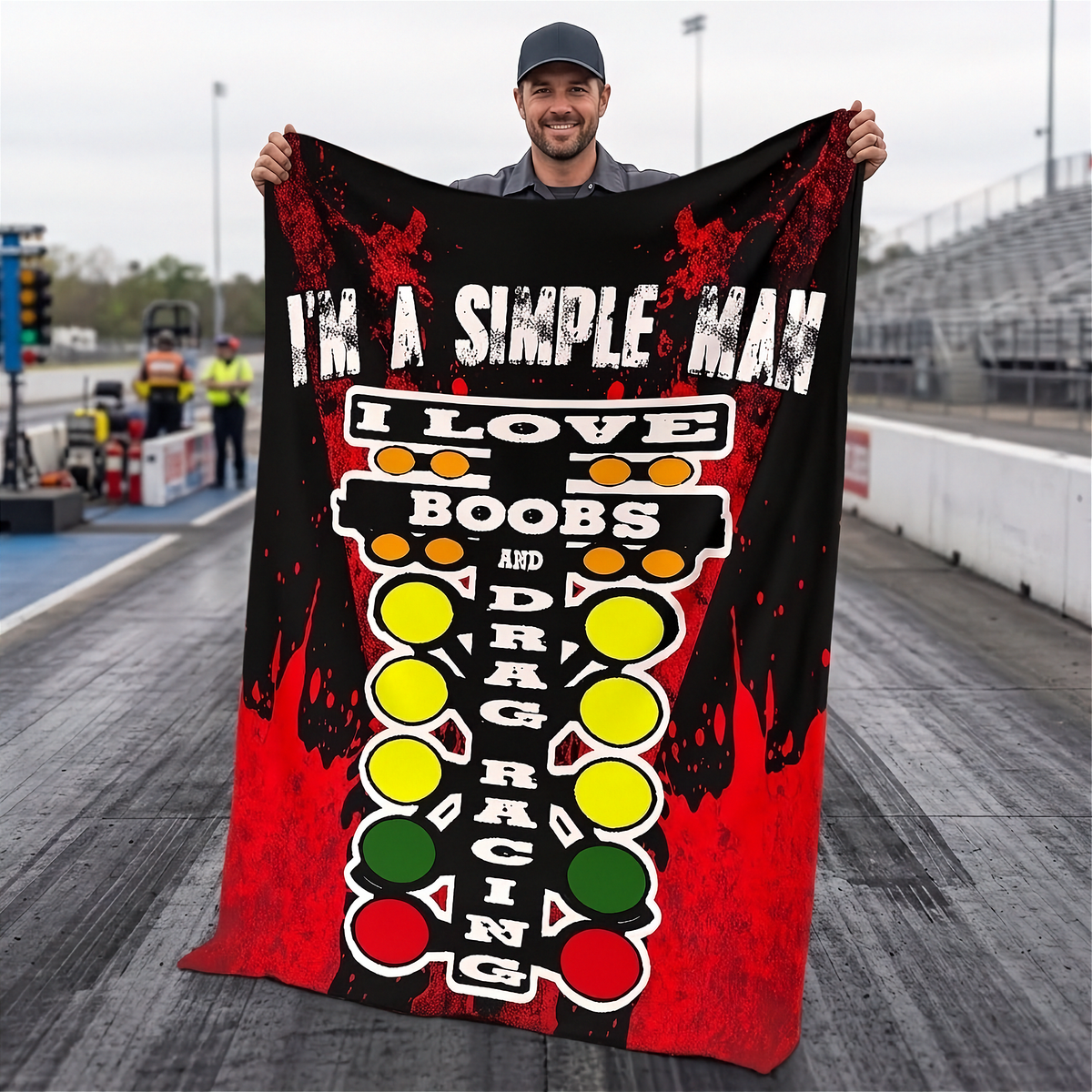 A man stands on a drag racing track holding up a large black and red blanket with a graphic design. The design features the text 'I'M A SIMPLE MAN I LOVE BOOBS AND DRAG RACING' above a vertical drag racing light pattern in red, yellow, and green.