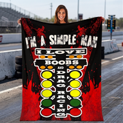 A woman stands on a racetrack holding up a large printed blanket or poster. The product has a black background with red splatter graphics, white text reading 'I'M A SIMPLE MAN I LOVE BOOBS AND DRAG RACING,' and a vertical racing-style light sequence made of yellow, green, and red circles.