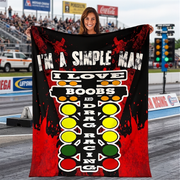 A woman stands on a racetrack holding up a large black and red blanket or towel with a bold graphic design. The design features the text 'I'M A SIMPLE MAN I LOVE BOOBS AND DRAG RACING' above a vertical sequence of traffic light-style circles in red, yellow, and green.