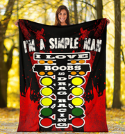 A woman stands outdoors in a wooded area holding up a large printed blanket or tapestry. The blanket has a black background with red splatter graphics, white text reading 'I'M A SIMPLE MAN I LOVE BOOBS AND DRAG RACING,' and a vertical racing-style light bar with yellow, green, and red circular buttons.