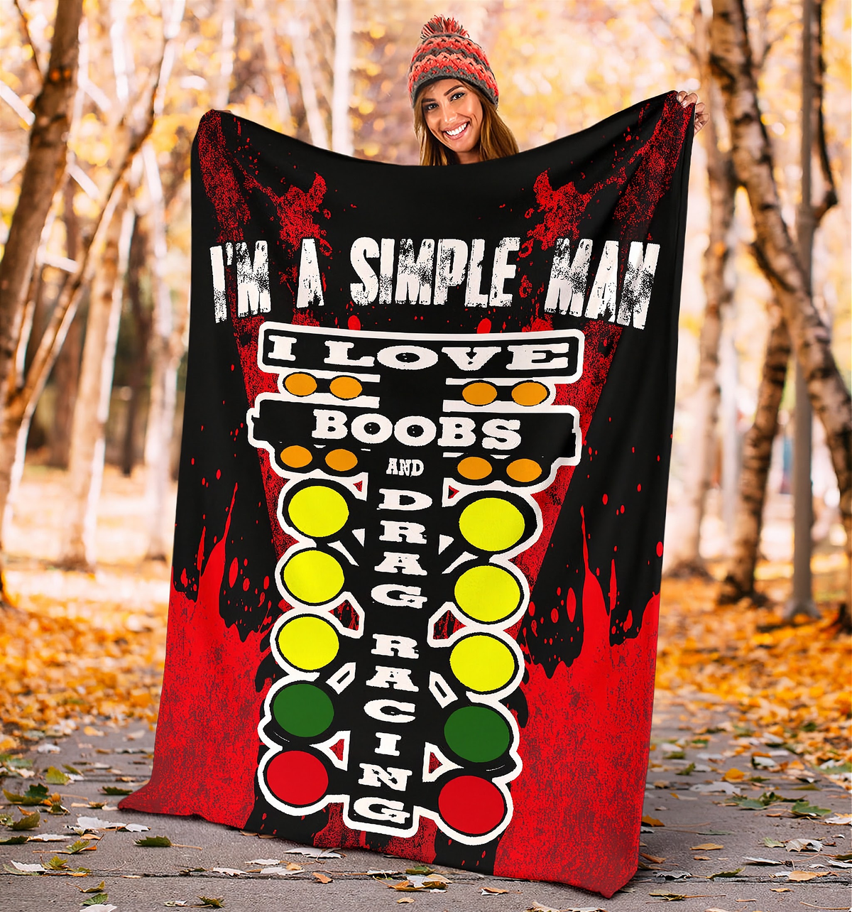 A woman wearing a knit beanie holds up a large printed blanket or poster outdoors in a wooded park setting. The product has a black and red distressed design with white text reading 'I'M A SIMPLE MAN' and a stylized racing track graphic in yellow, green, and red.