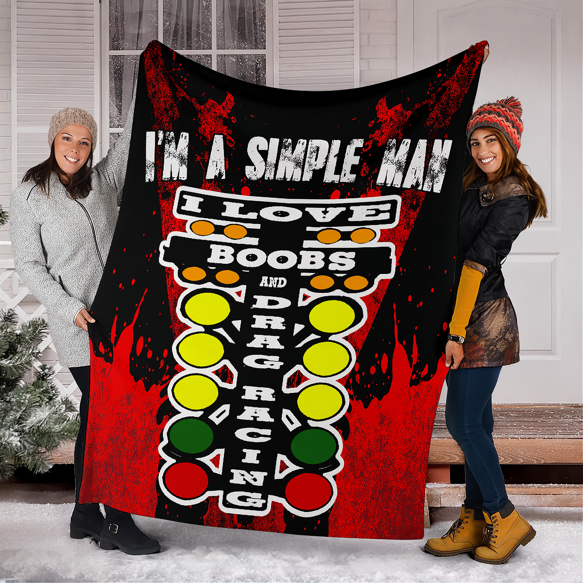 Two women standing in front of a house doorway holding up a large printed blanket or throw. The blanket has a black and red design with the text 'I'M A SIMPLE MAN' and a retro arcade-style graphic with yellow, green, and red buttons.