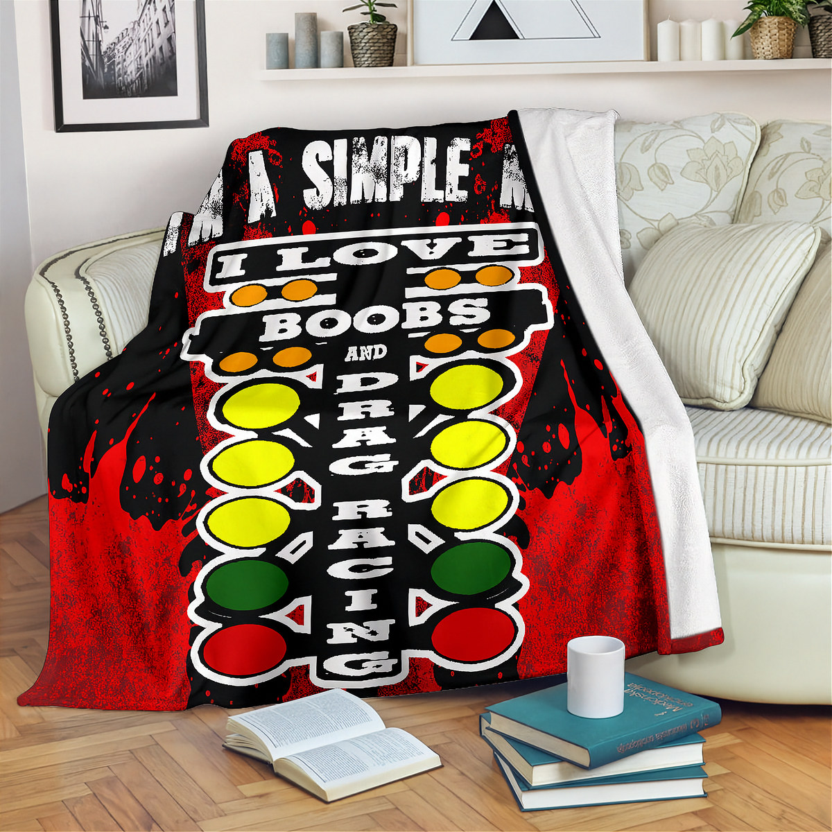 A red and black throw blanket draped over a cream sofa in a living room setting, featuring a graphic design with the words 'I LOVE BOOBS AND DRAG RACING' and circular traffic-light style icons in yellow, green, and red.