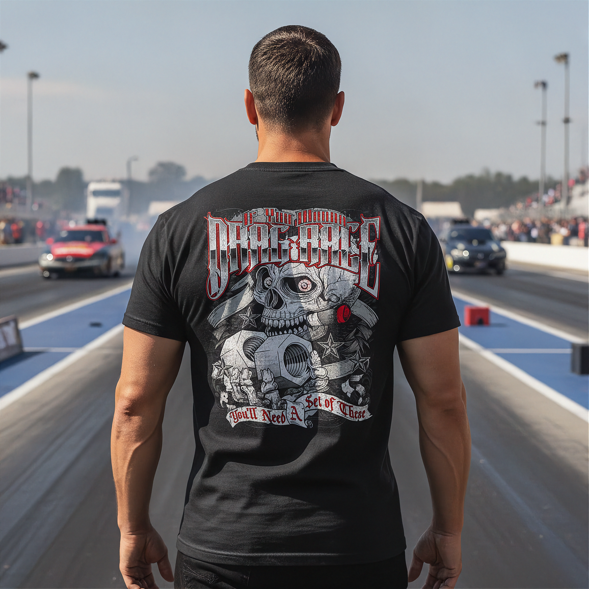A man standing on a drag strip wearing a black t-shirt with a large back graphic that reads 'if you want to Drag Race' and features a detailed skull and engine design.