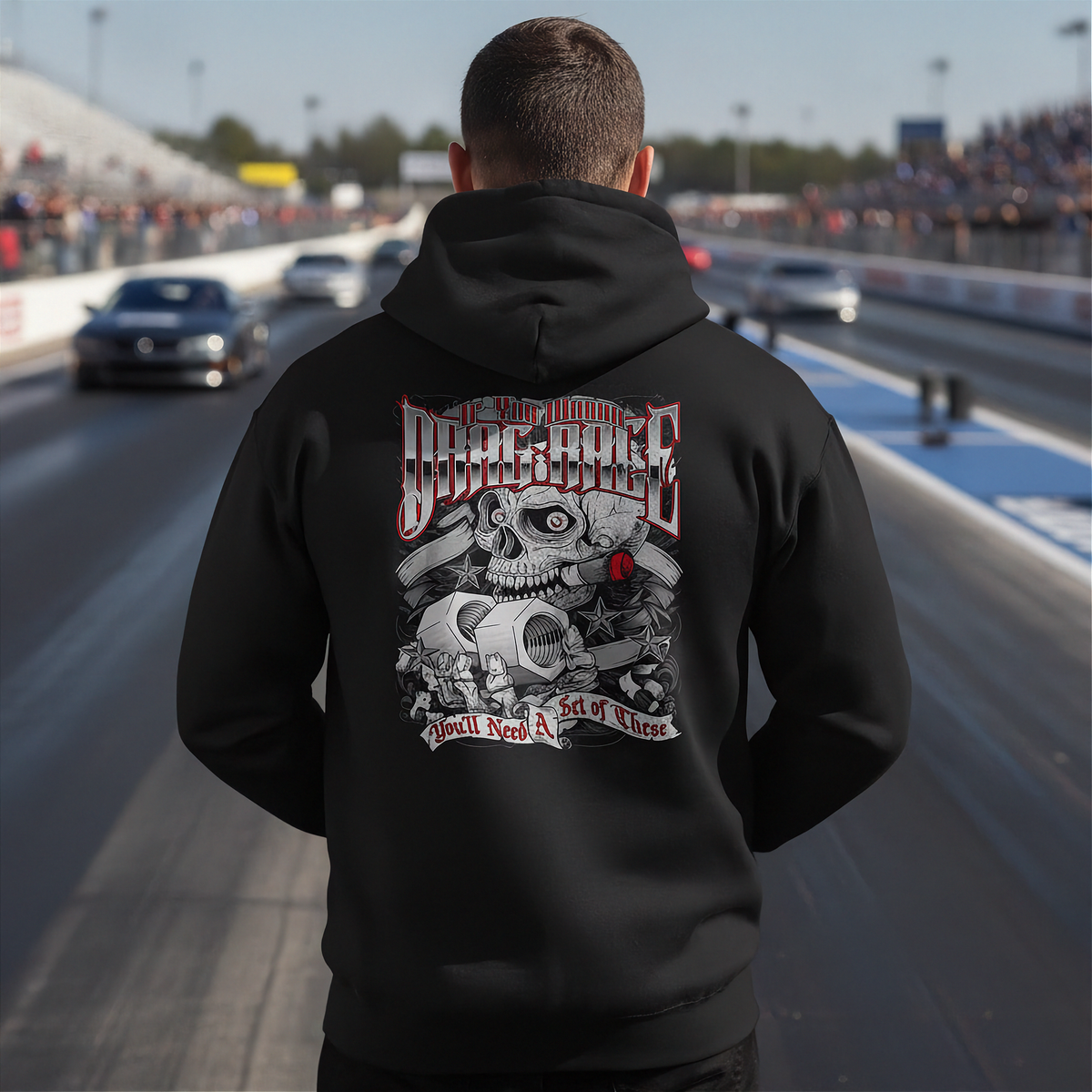 A man standing on a drag racing track, viewed from behind, wearing a black hoodie with a large graphic print on the back. The graphic features a skull, crossed bones, a red rose, and the text 'Drag Race' and 'You'll Need A Set of Vices'.