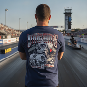 A man stands on a drag racing track wearing a navy blue t-shirt with a large back graphic featuring a skull, gears, and the text 'Drag race' and 'You Need A'. The background shows a blurred racetrack, grandstands, and a tower.