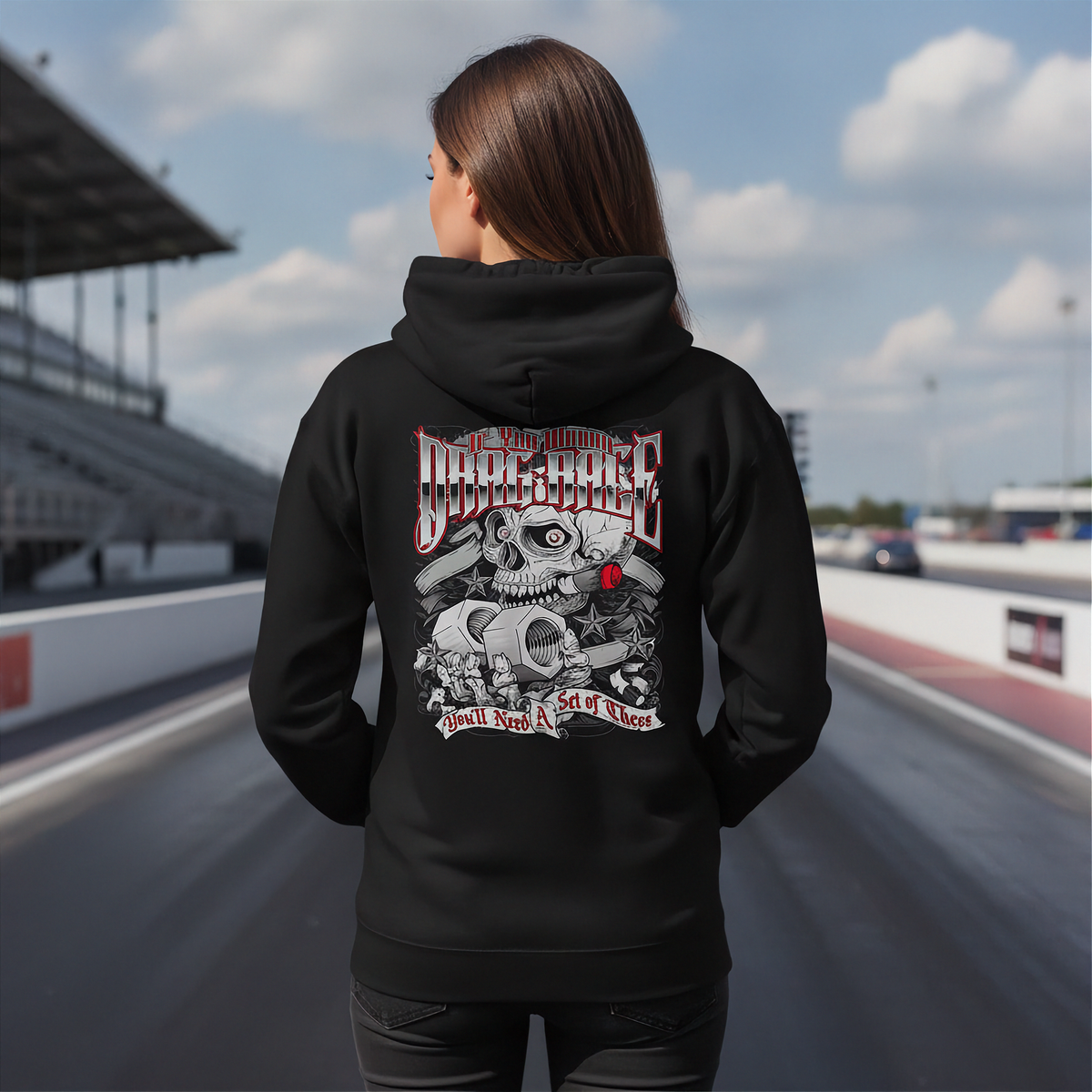 A woman viewed from behind wearing a black pullover hoodie with a large graphic print on the back, standing on a racetrack with grandstands and a cloudy sky in the background.
