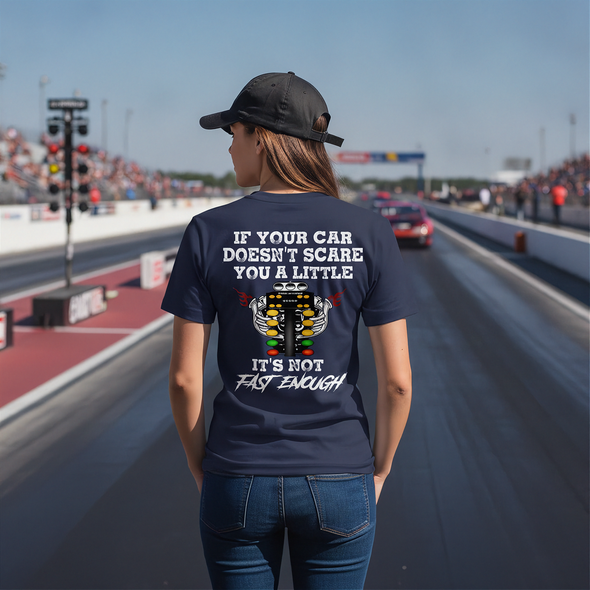 A woman stands on a racetrack wearing a navy blue t-shirt with a humorous racing graphic on the back. The graphic features a white engine illustration and the text 'IF YOUR CAR DOESN'T SCARE YOU A LITTLE IT'S NOT FAST ENOUGH'.