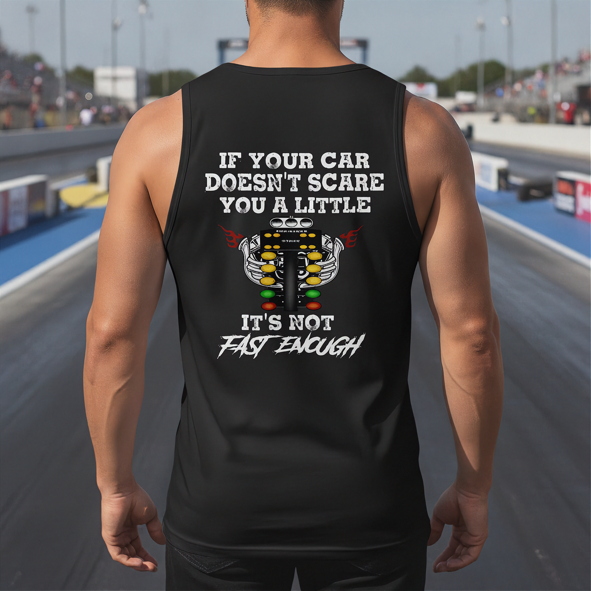 A man stands on a racetrack wearing a black tank top with a humorous racing graphic on the back. The graphic features a white engine illustration and the text 'IF YOUR CAR DOESN'T SCARE YOU A LITTLE IT'S NOT FAST ENOUGH'.