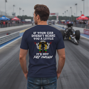 A man stands on a racetrack wearing a navy t-shirt with a humorous racing graphic on the back. The graphic features a white engine illustration and the text 'IF YOUR CAR DOESN'T SCARE YOU A LITTLE IT'S NOT FAST ENOUGH'.