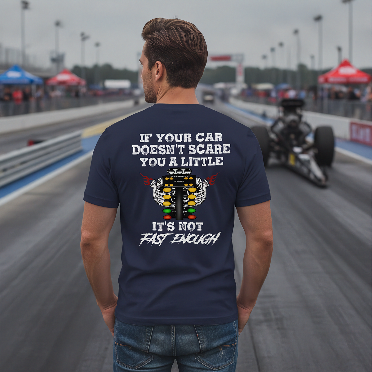 A man stands on a racetrack wearing a navy t-shirt with a humorous racing graphic on the back. The graphic features a white engine illustration and the text 'IF YOUR CAR DOESN'T SCARE YOU A LITTLE IT'S NOT FAST ENOUGH'.