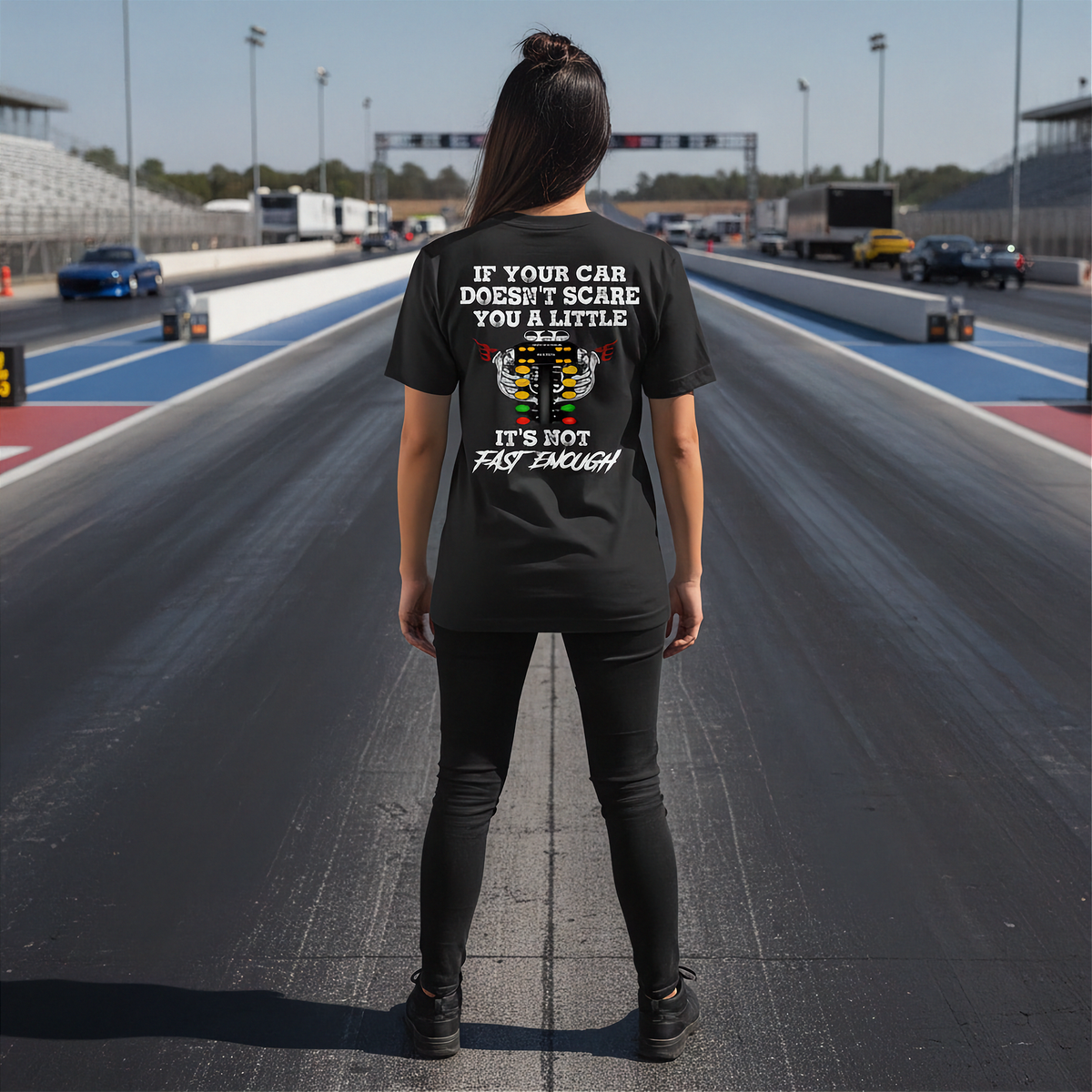 A woman stands on a racetrack wearing a black t-shirt with a humorous racing graphic on the back. The graphic features a white engine illustration and the text 'IF YOUR CAR DOESN'T SCARE YOU A LITTLE IT'S NOT FAST ENOUGH'.