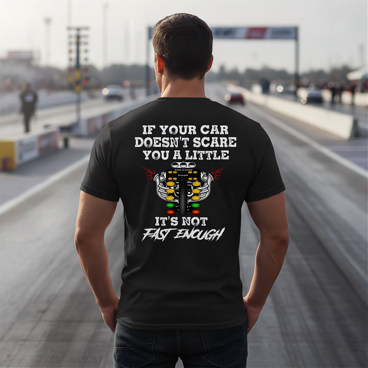 A man stands on a racetrack wearing a black t-shirt with a humorous racing graphic on the back. The graphic features a white engine illustration and the text 'IF YOUR CAR DOESN'T SCARE YOU A LITTLE IT'S NOT FAST ENOUGH'.