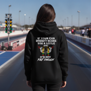 A man stands on a racetrack wearing a black hoodie with a humorous racing graphic on the back. The graphic features a white engine illustration and the text 'IF YOUR CAR DOESN'T SCARE YOU A LITTLE IT'S NOT FAST ENOUGH'.