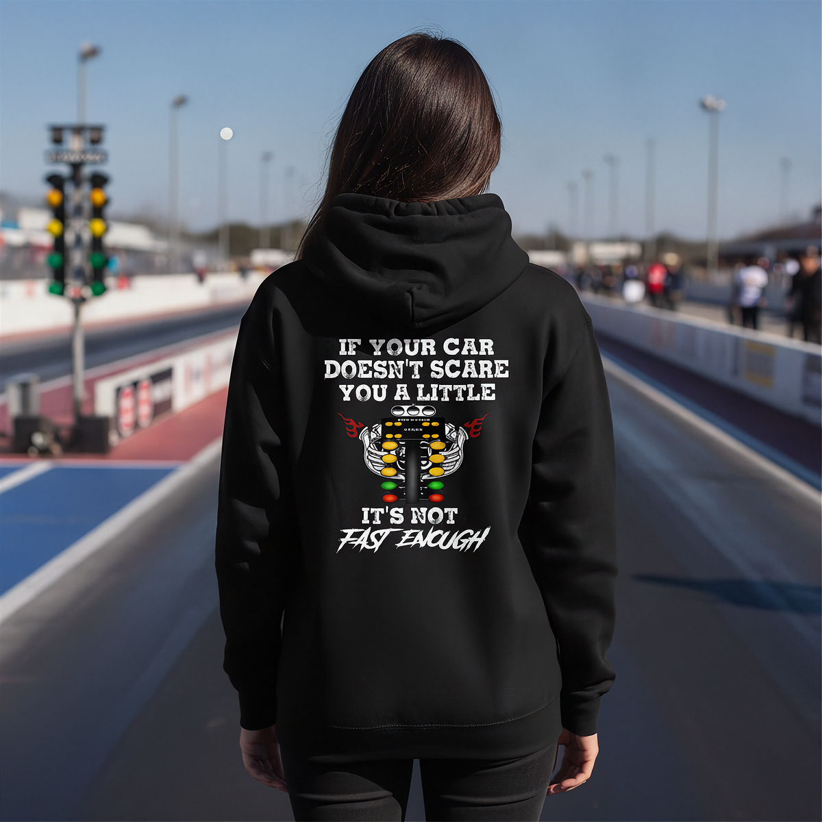 A man stands on a racetrack wearing a black hoodie with a humorous racing graphic on the back. The graphic features a white engine illustration and the text 'IF YOUR CAR DOESN'T SCARE YOU A LITTLE IT'S NOT FAST ENOUGH'.