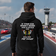 A man stands on a racetrack wearing a black hoodie with a humorous racing graphic on the back. The graphic features a white engine illustration and the text 'IF YOUR CAR DOESN'T SCARE YOU A LITTLE IT'S NOT FAST ENOUGH'.