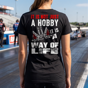 A woman at a drag race track wearing a black shirt with 'It Is Not Just A Hobby IT'S MY WAY OF LIFE'